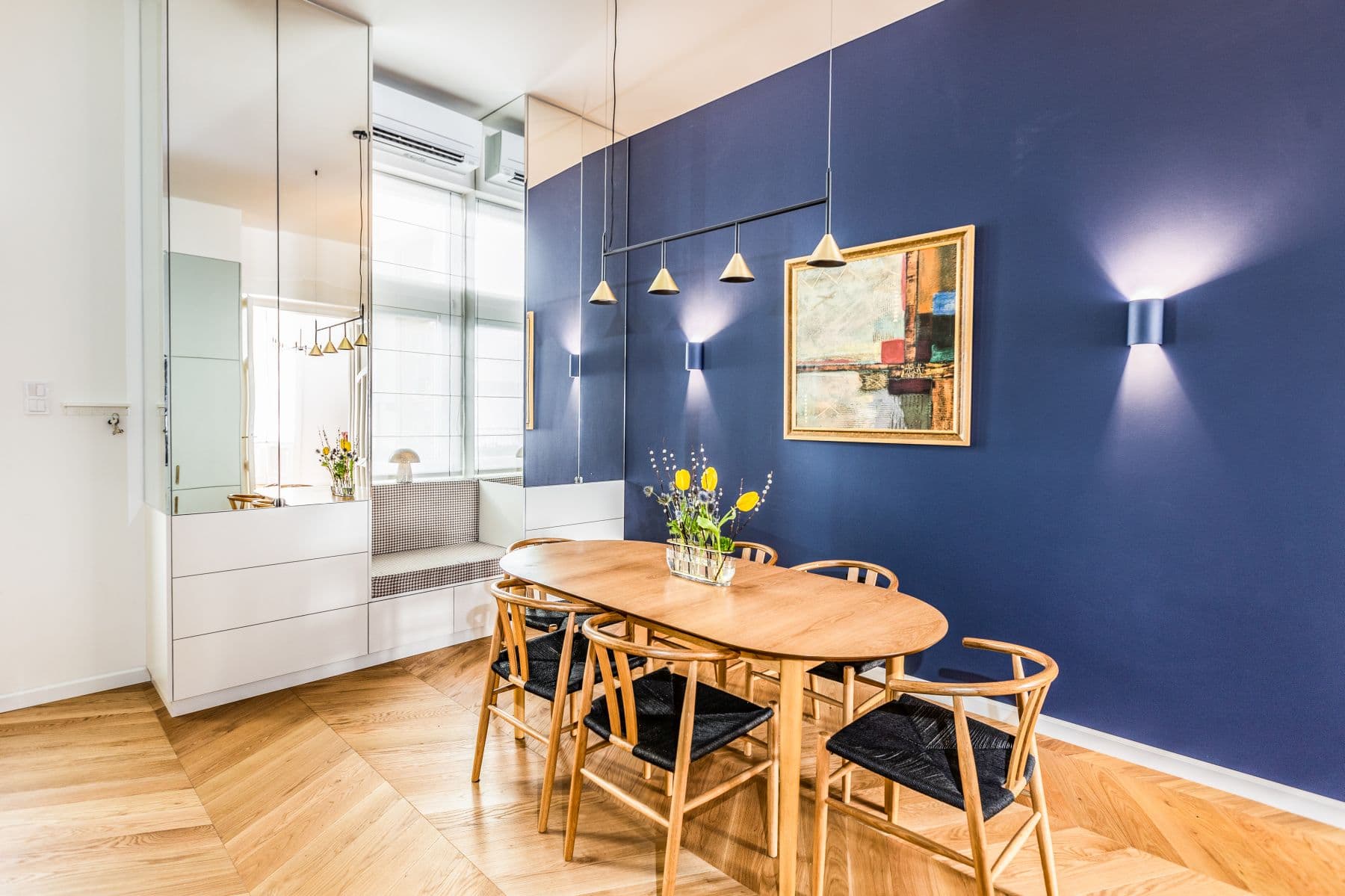Modern dining room with an oval wooden table, blue wall, and gold framed abstract painting.