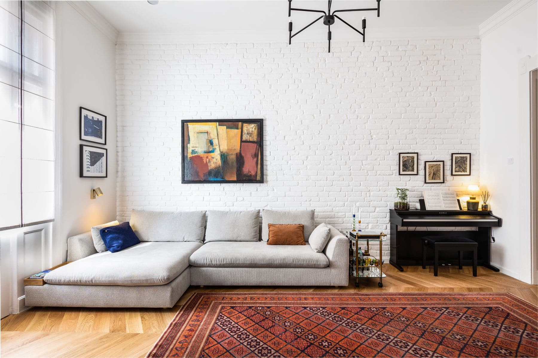 Modern living room with a gray sectional, white brick wall, abstract art, and piano.