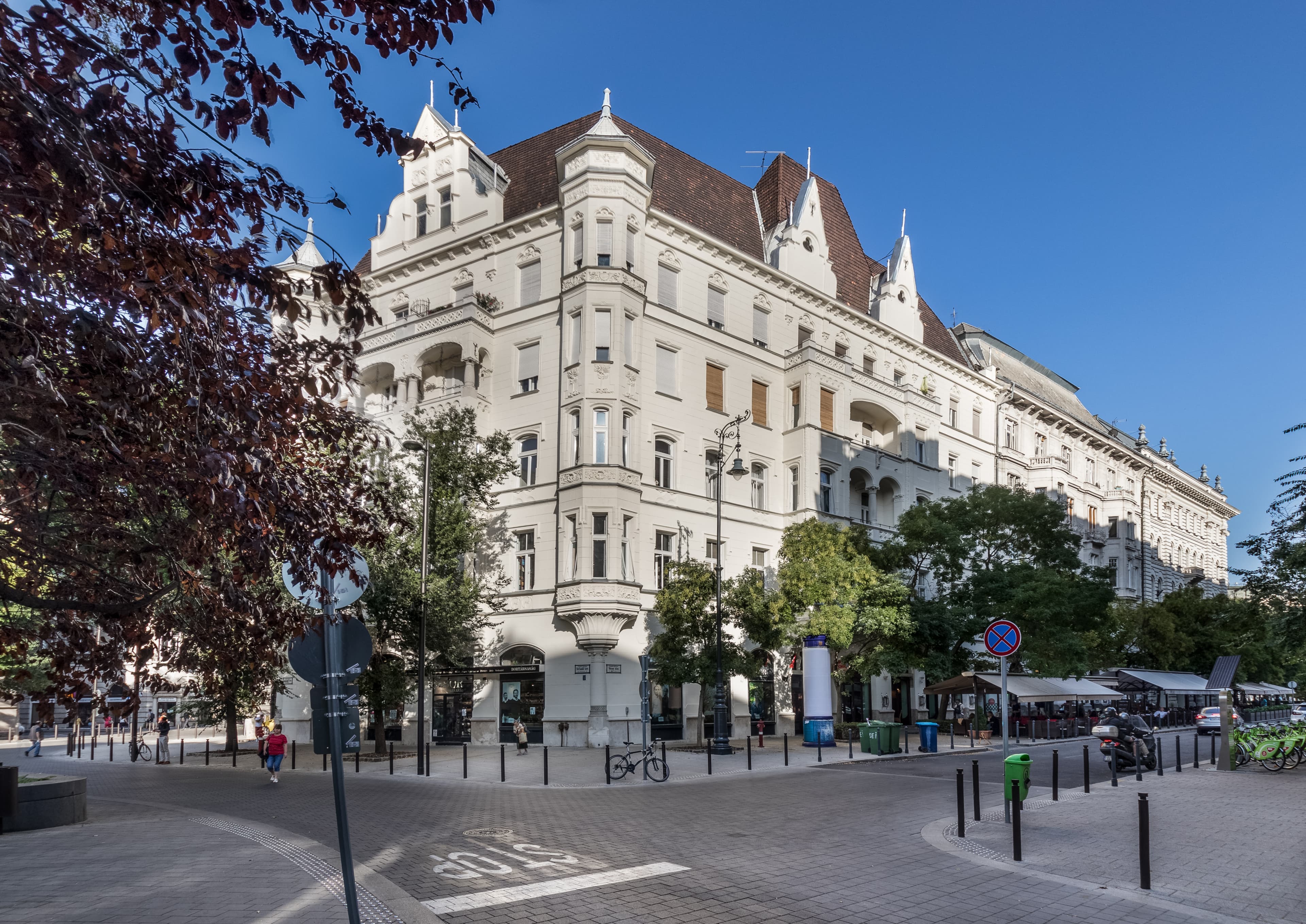 Ornate white building on a sunny European street corner with trees and blue sky.